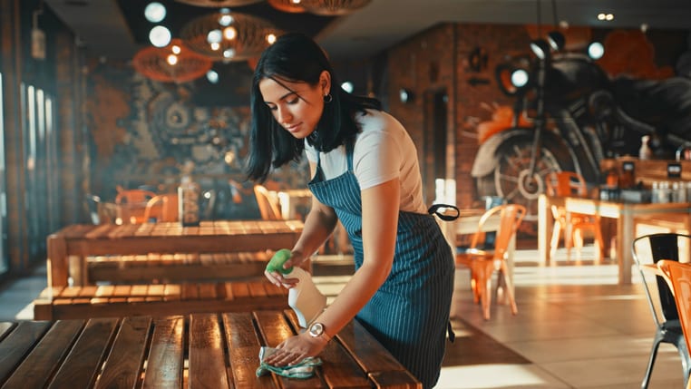 A woman in an apron cleaning a table in a restaurant.