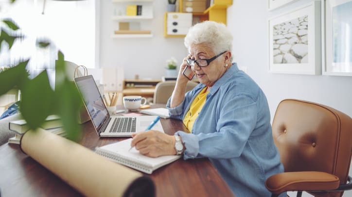 An elderly woman sitting at a desk and talking on the phone.