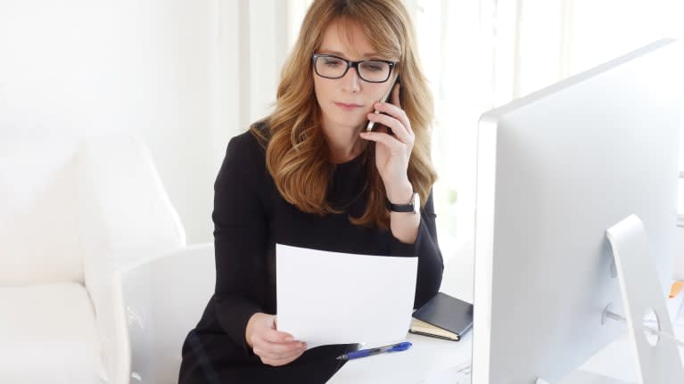 A woman in glasses is talking on the phone while sitting at her desk.