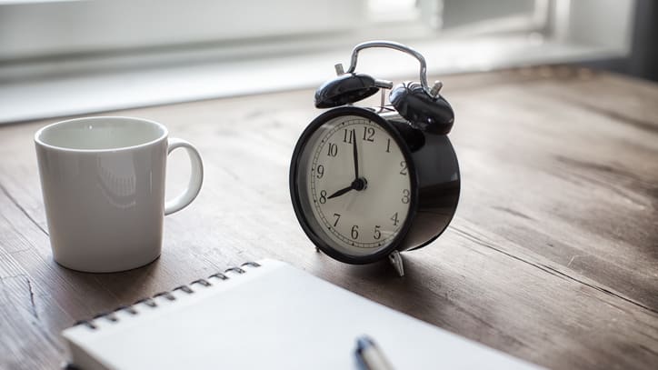 A black alarm clock next to a notebook and a cup of coffee.