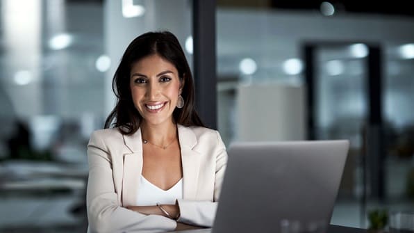 A smiling business woman with her arms crossed in front of a laptop.