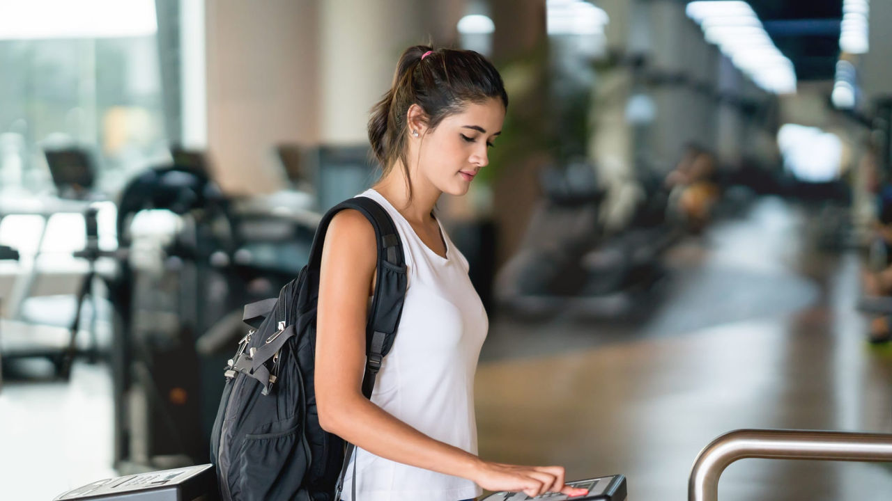 A woman with a backpack standing at a metal gate in a gym.