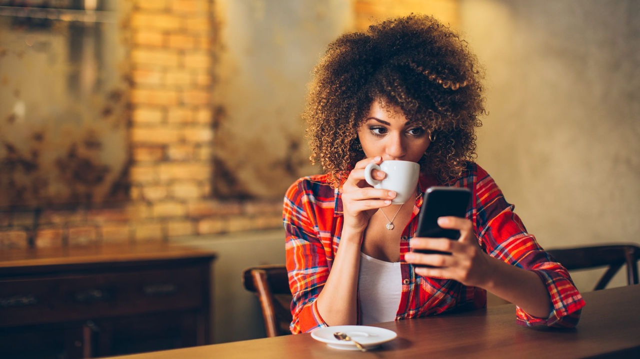 A woman with afro hair drinking coffee while looking at her phone.