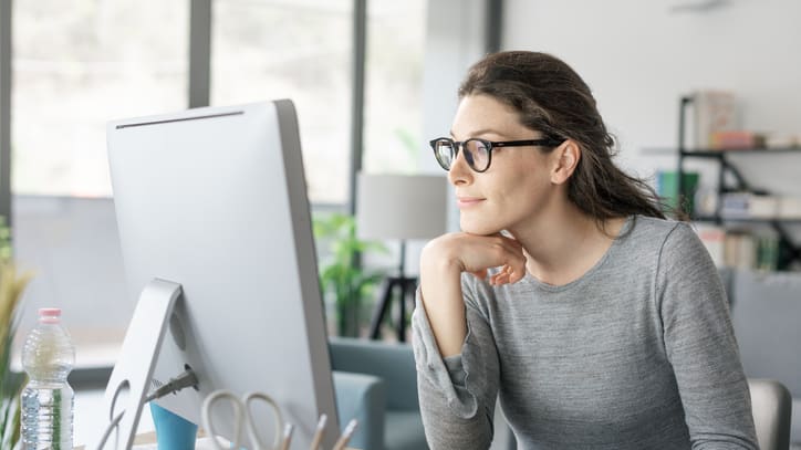 A woman in glasses is sitting at a desk looking at her computer.