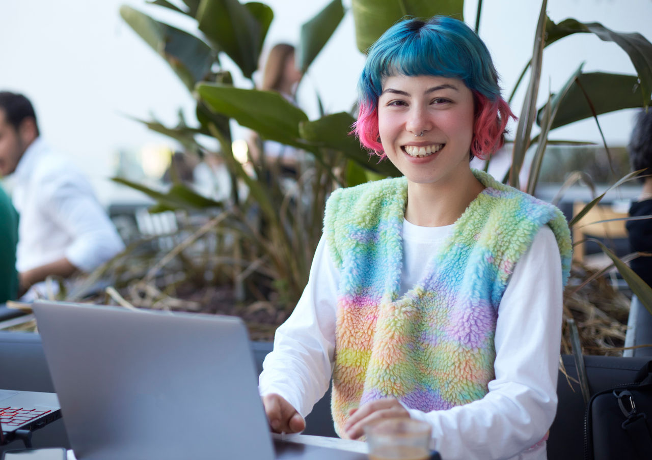 Smiling young woman with colorful hair working with laptop computer in cafe.