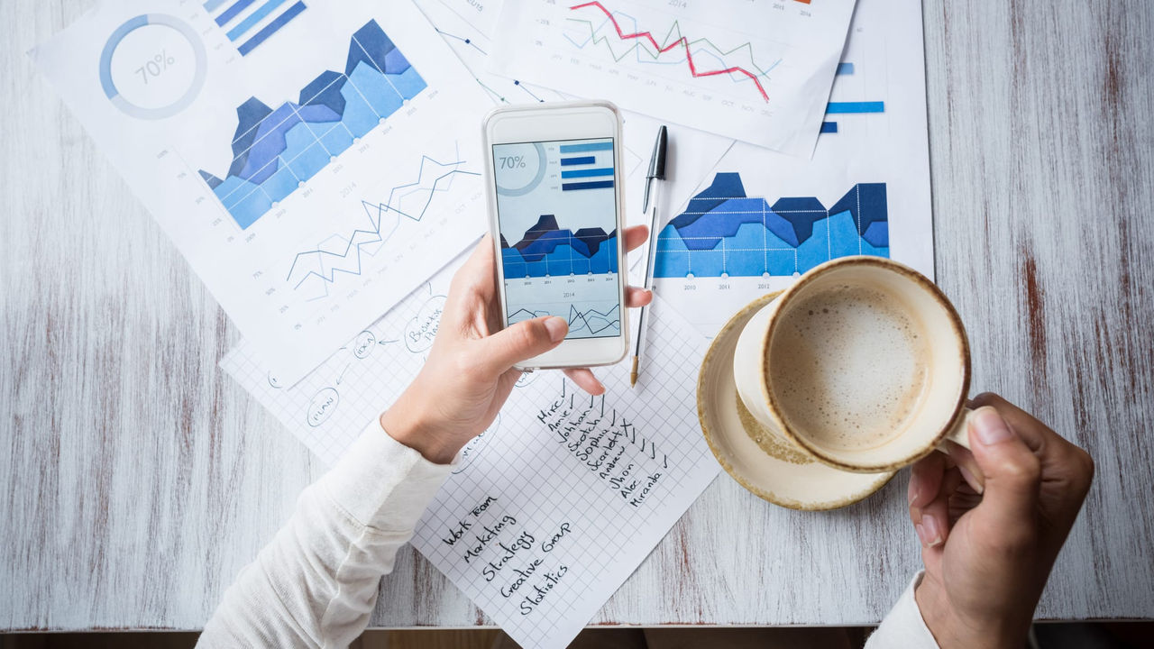 A woman is holding a smartphone while looking at graphs on a table.