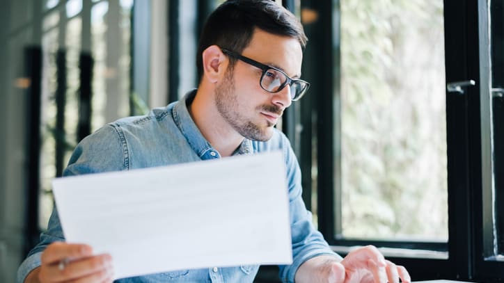 A man in glasses is looking at a piece of paper while sitting at a desk.