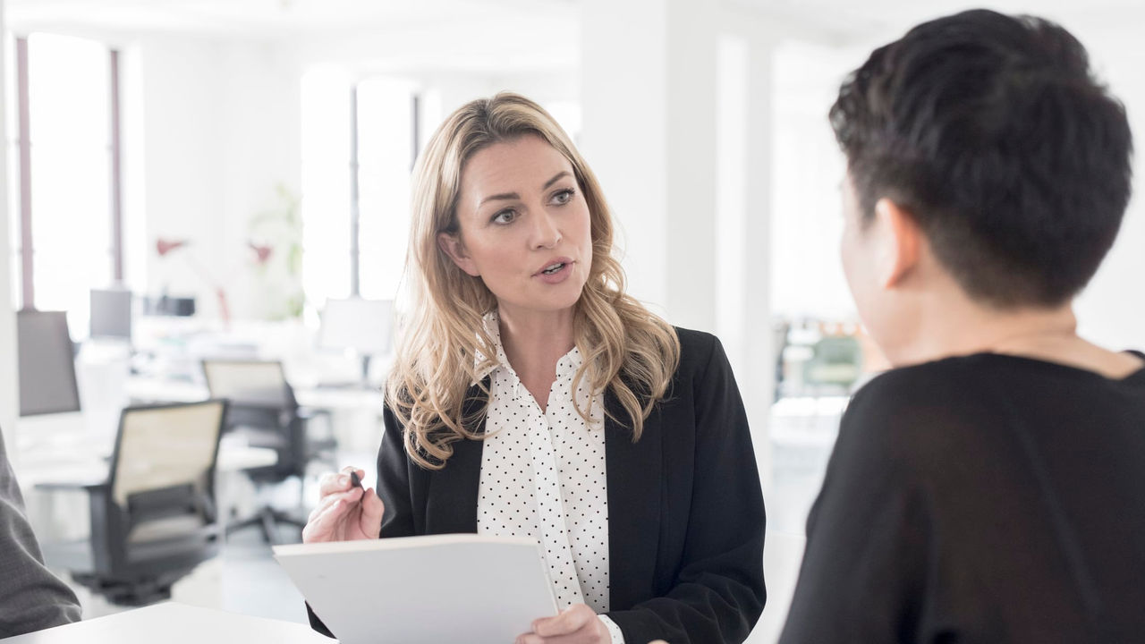 A businesswoman holding a stack of papers is talking to an employee in an office.