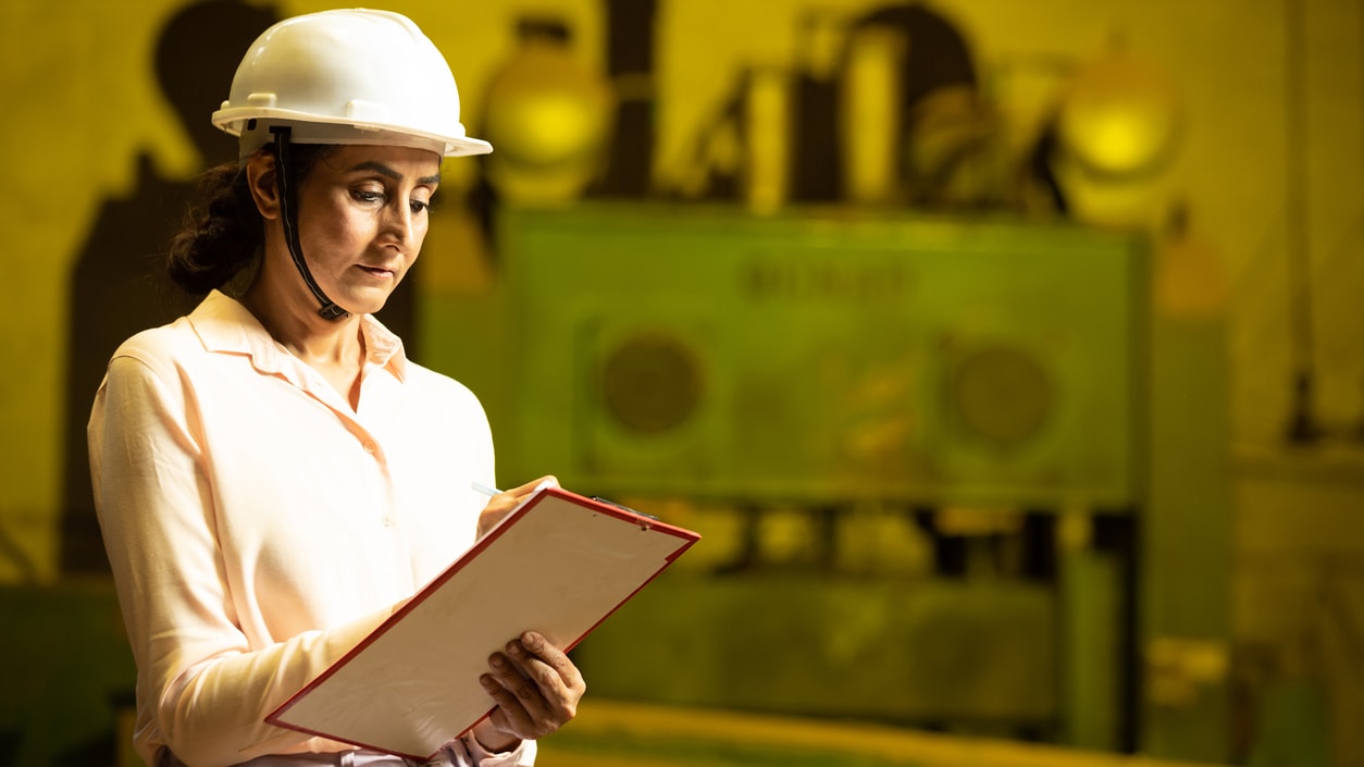A woman wearing a hard hat is holding a clipboard in front of a factory.