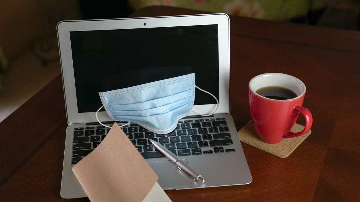 A laptop with a medical mask and a cup of coffee on a wooden table.