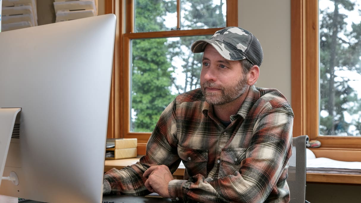 A man in a plaid shirt sitting at a desk with a computer.