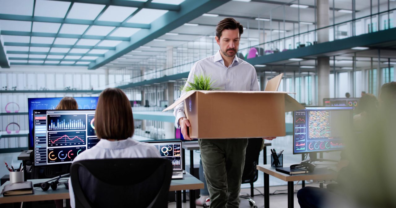 A man carrying a cardboard box walks through an office after being laid off. 