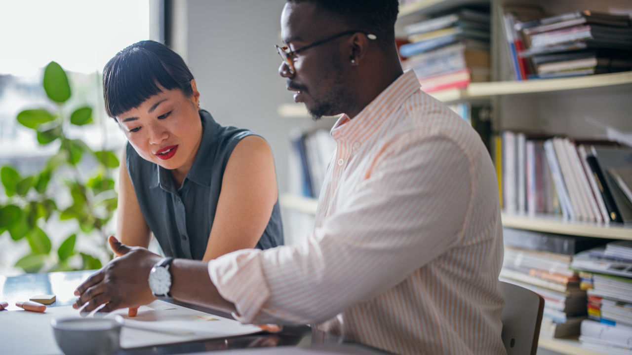 A serious African-American businessman with glasses explaining something to his beautiful Japanese coworker while they are sitting at the desk. (mentorship concept)