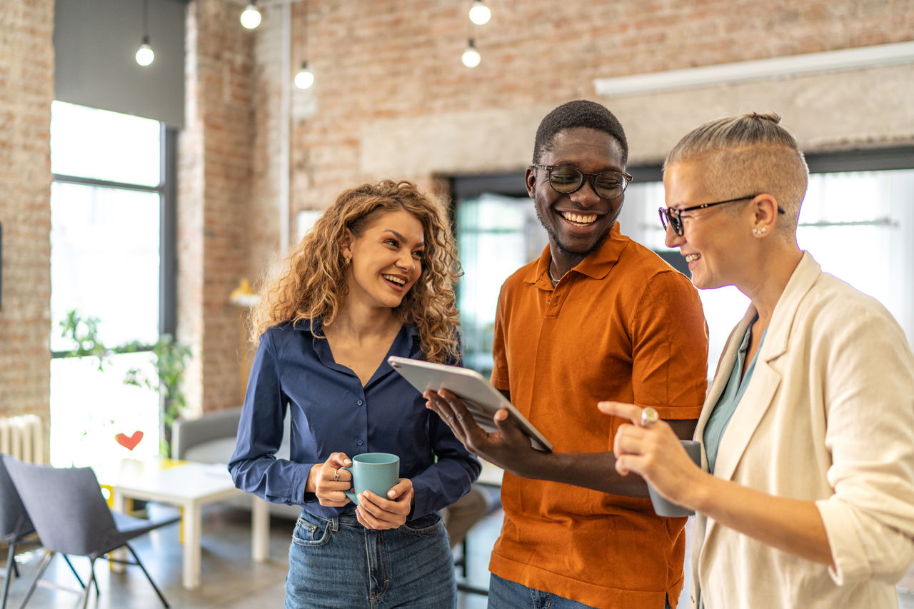 Diverse business professionals smiling and discussing work while looking at a digital tablet, fostering collaboration and teamwork in a creative, open-plan office environment