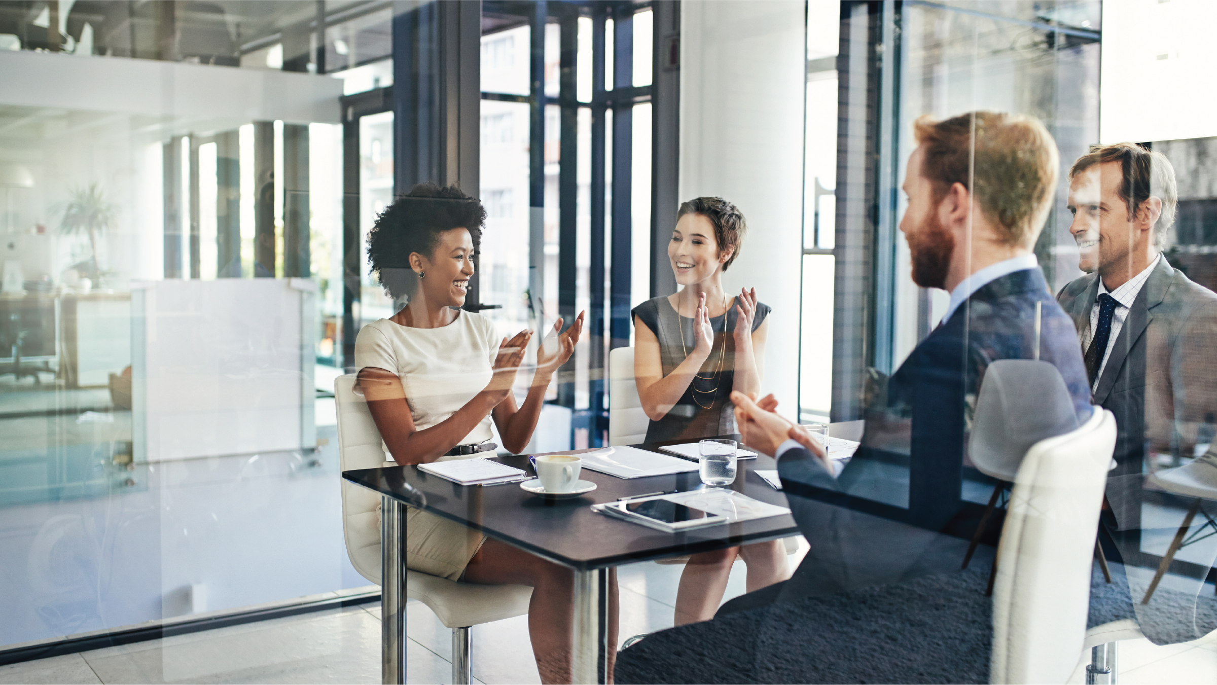 men and women clap while meeting at a table in a modern glass office