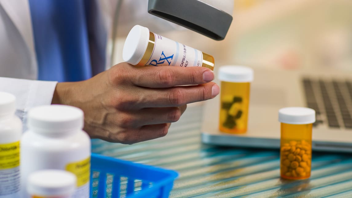 A pharmacist is holding a bottle of medicine in front of a laptop.