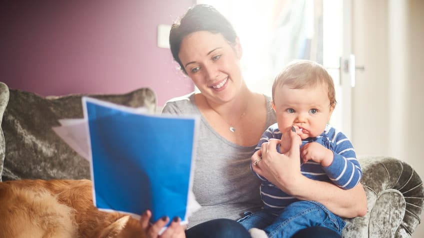 A woman reading a letter to her baby while sitting on a couch with a dog.