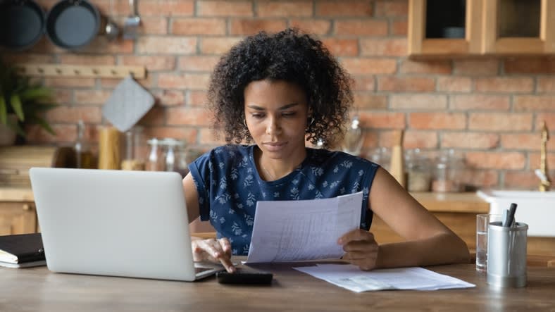 A woman is sitting at a kitchen table with a laptop and papers.