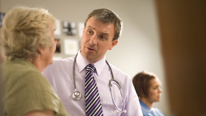 A male doctor talking to an elderly woman in a waiting room.