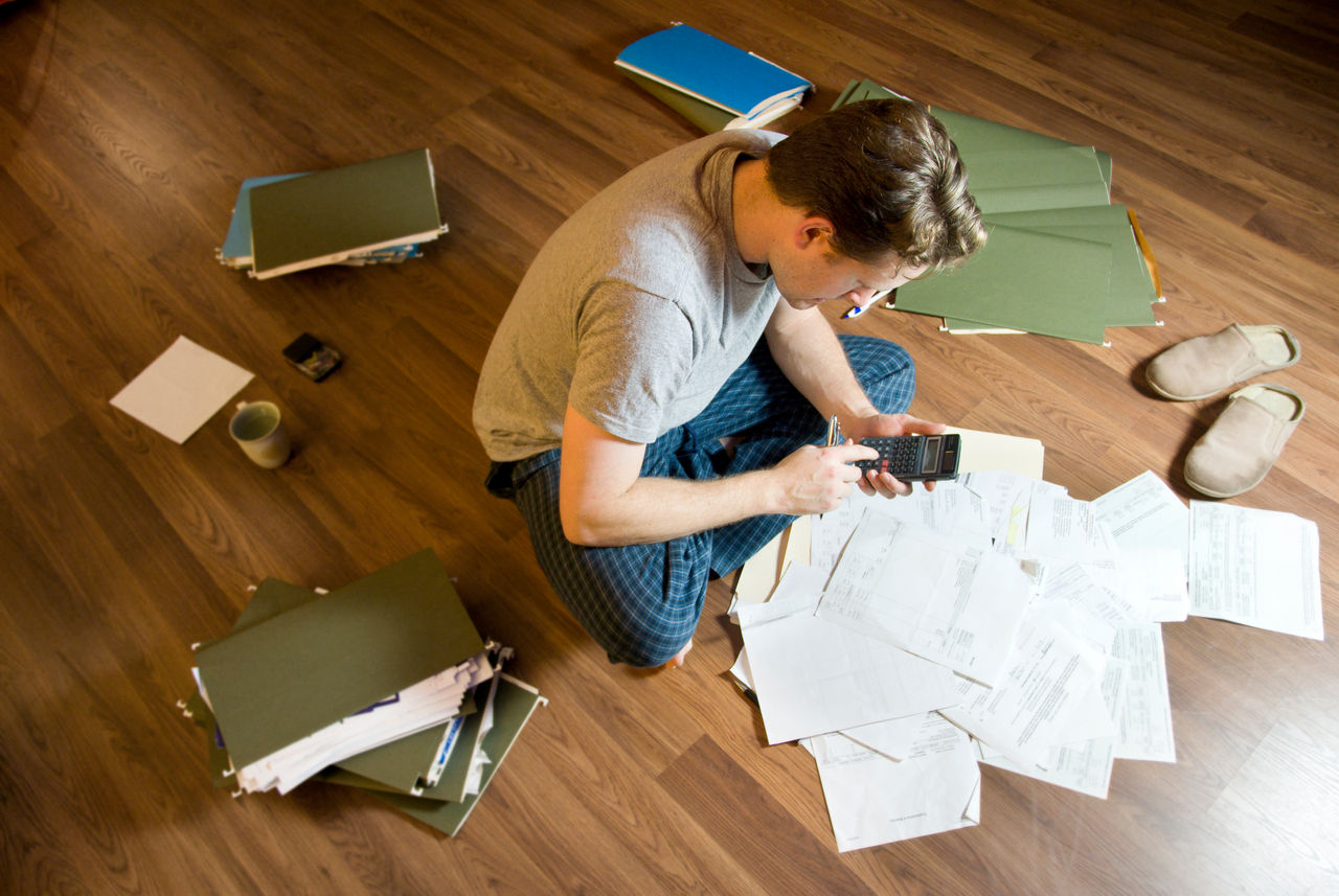 Man sits on floor surrounded by files and paperwork, holding calculator