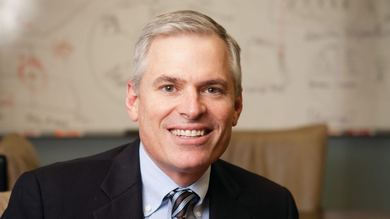 A man in a suit and tie smiling in front of a whiteboard.