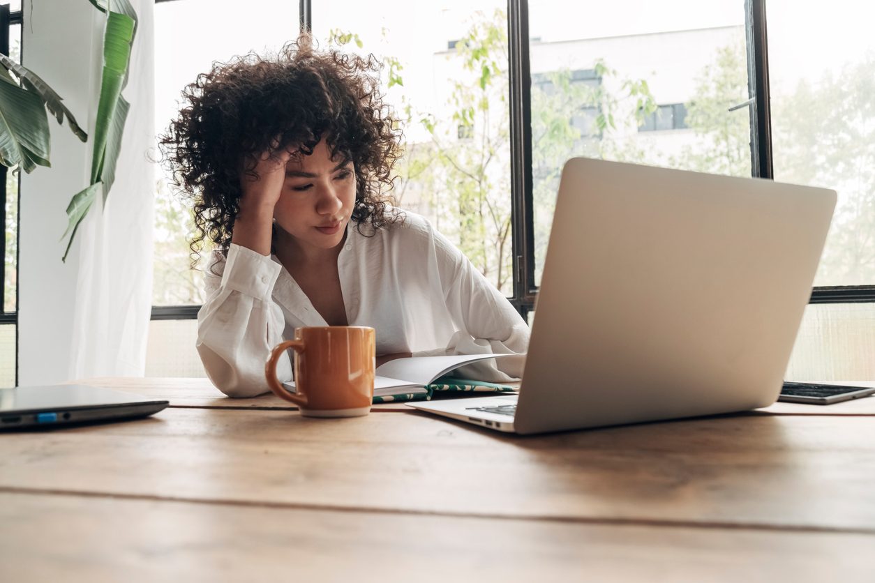 Woman sitting and thinking in front of a desk with a mug and a laptop.