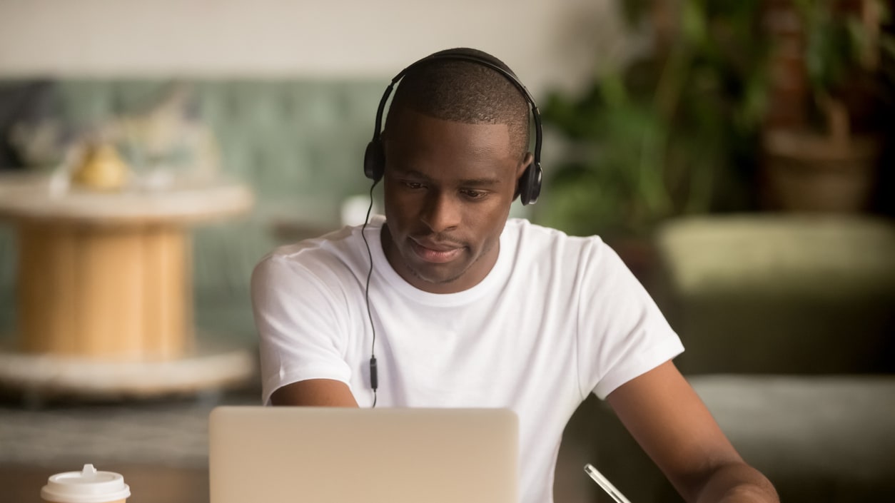 A man wearing a headset is working on a laptop in a coffee shop.