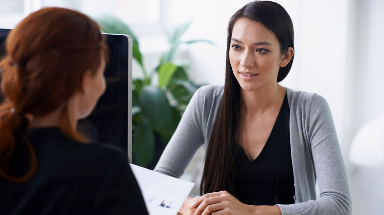 A woman is talking to another woman at a desk.